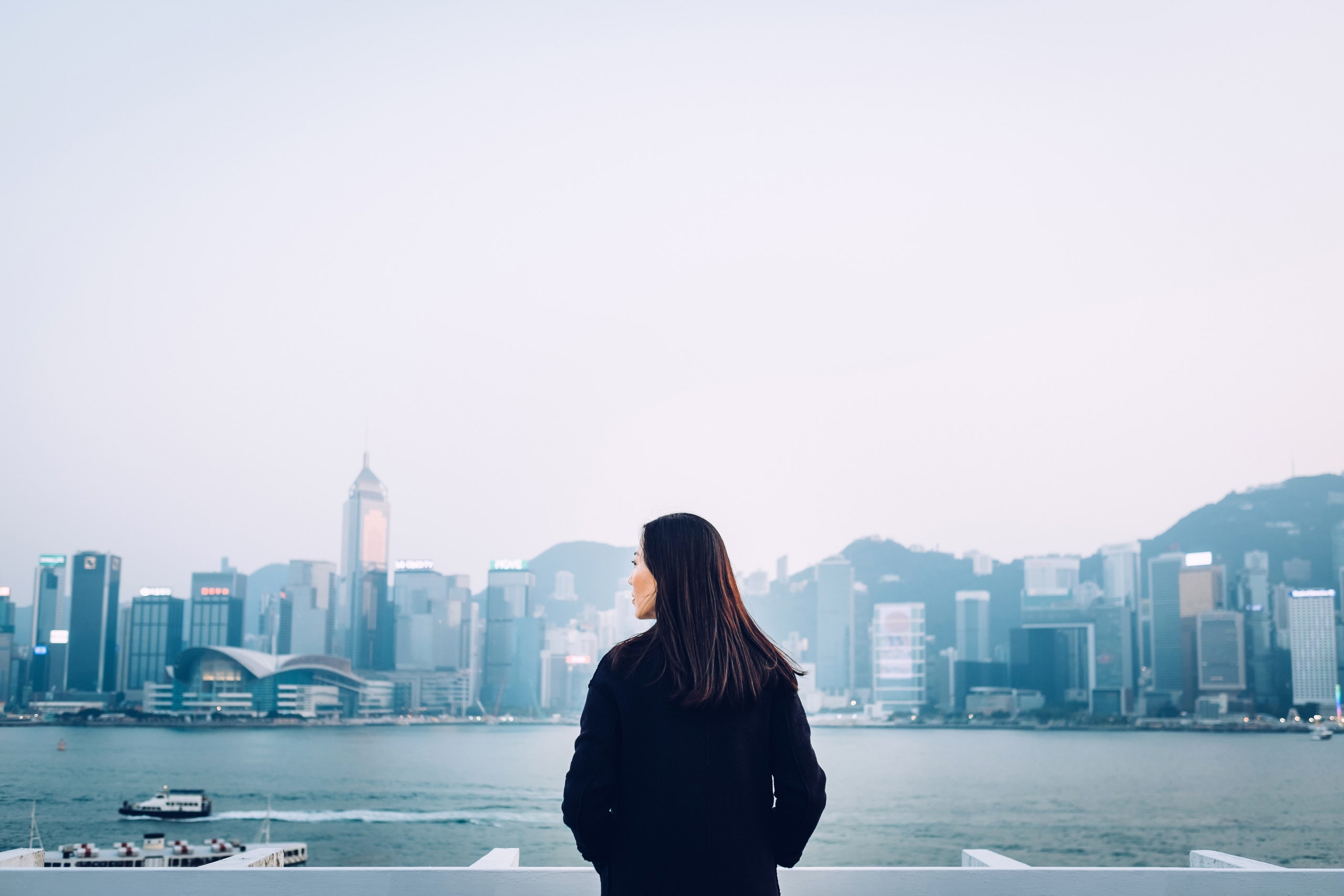 Vista traseira de uma jovem parada à beira-mar de Victoria Harbour, com vista para o horizonte da cidade de Hong Kong