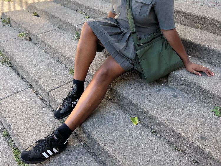 Influente Imani Randolph em tênis negros adidas samba com uma saia cinza e um saco verde de Crossbodi.