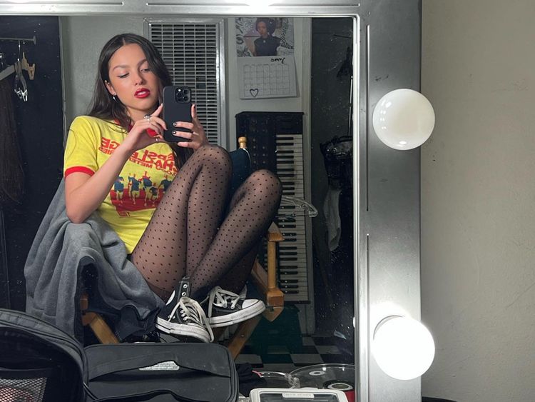 Olivia Rodrigo em uma camiseta gráfica, calças justas com um padrão de ponto e converse faz uma selfie no espelho.