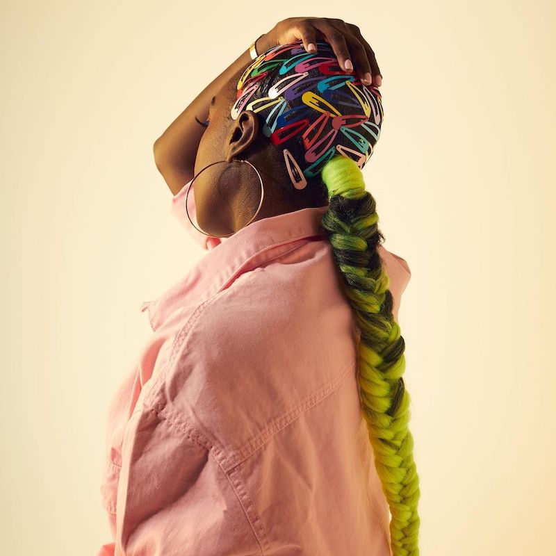Parte de trás da cabeça de uma mulher com clipes de arco-íris na raiz e um longo rabo de cavalo trançado com pontas amarelas
