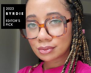 Editor de Byrdie Iden Stewart com lábios brilhantes, óculos de tartaruga e maquiagem brilhante
