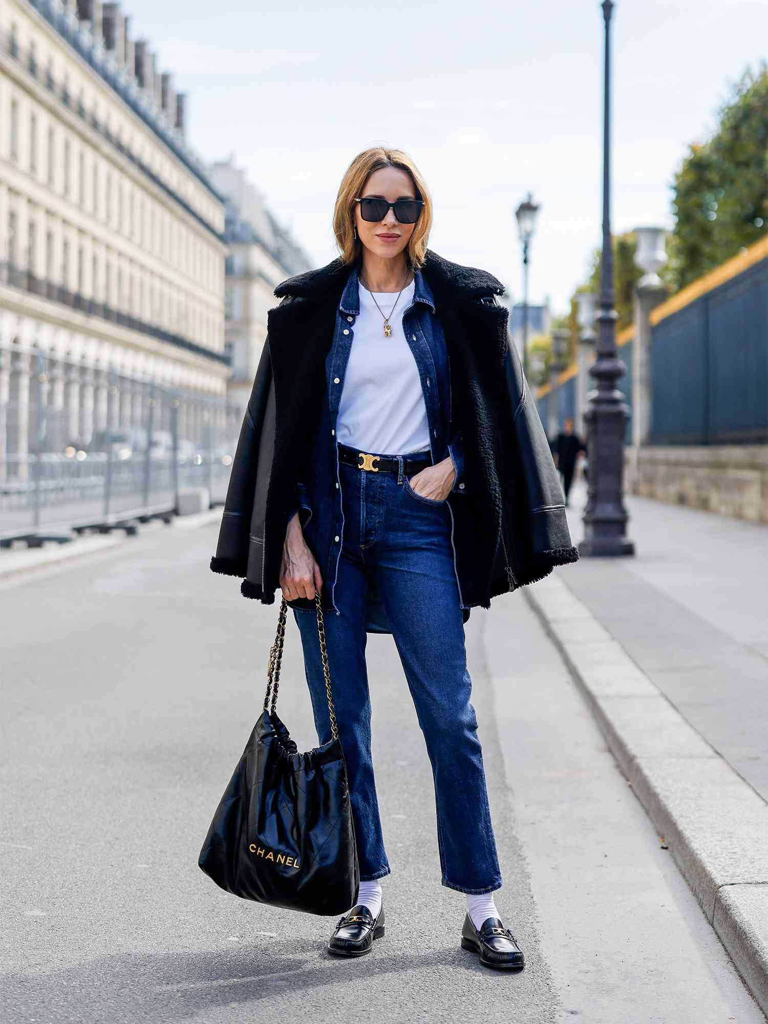 Uma mulher de jeans, jeans, uma camiseta branca, uma jaqueta de couro, uma grande bolsa Chanel, mocassins, meias e óculos de sol.