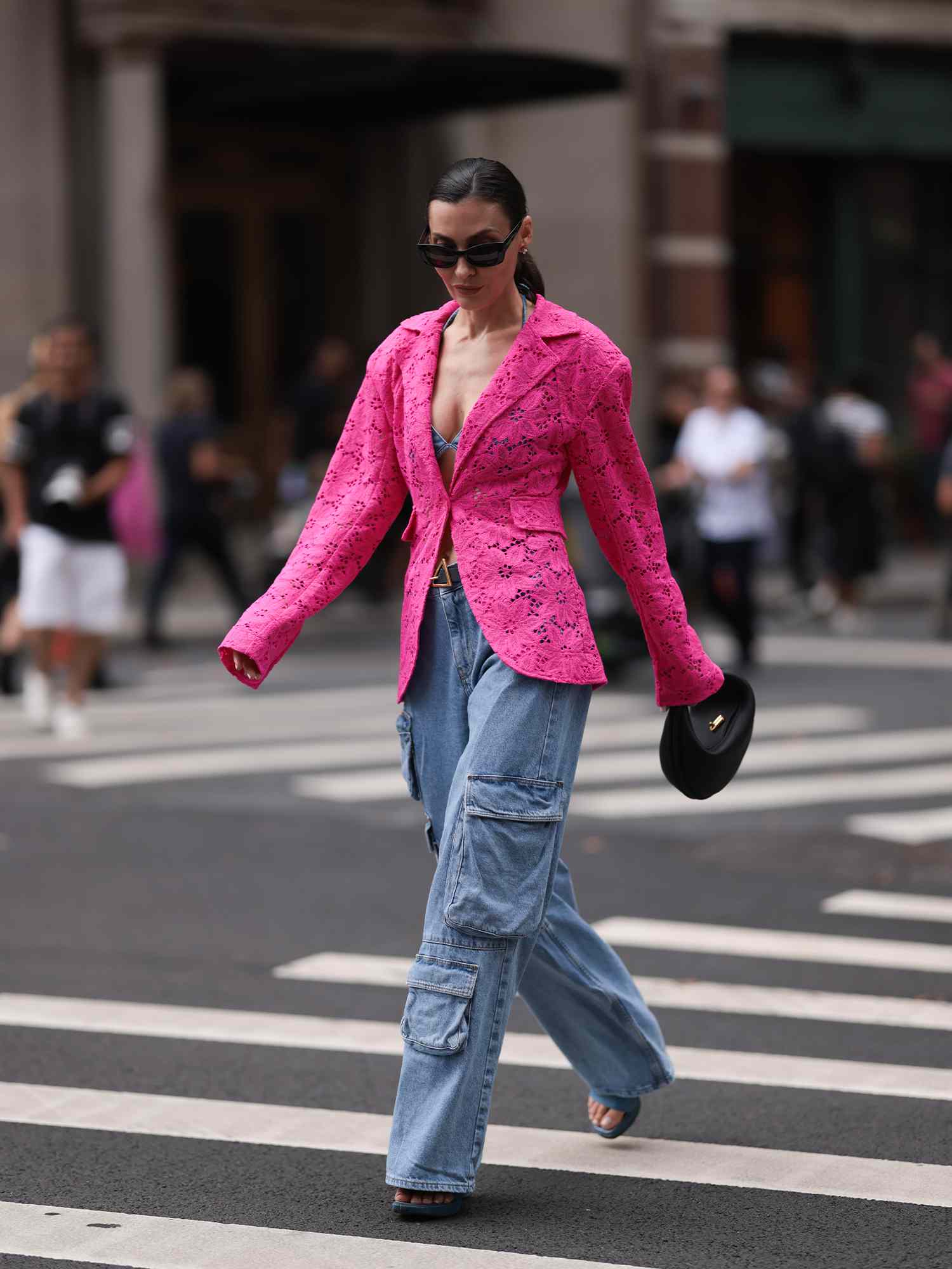 Uma mulher em um blazer de malha rosa, jeans-cargo, sapatos abertos, bolsa preta e óculos de sol