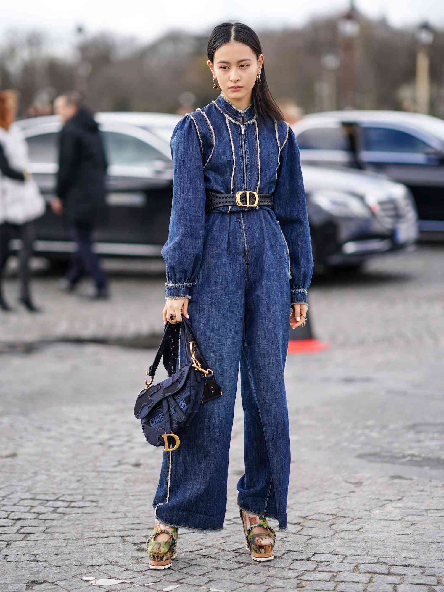 Lala Takahashi em um macacão de jeans azul com um cinto Dior, uma bolsa e sapatos em uma plataforma