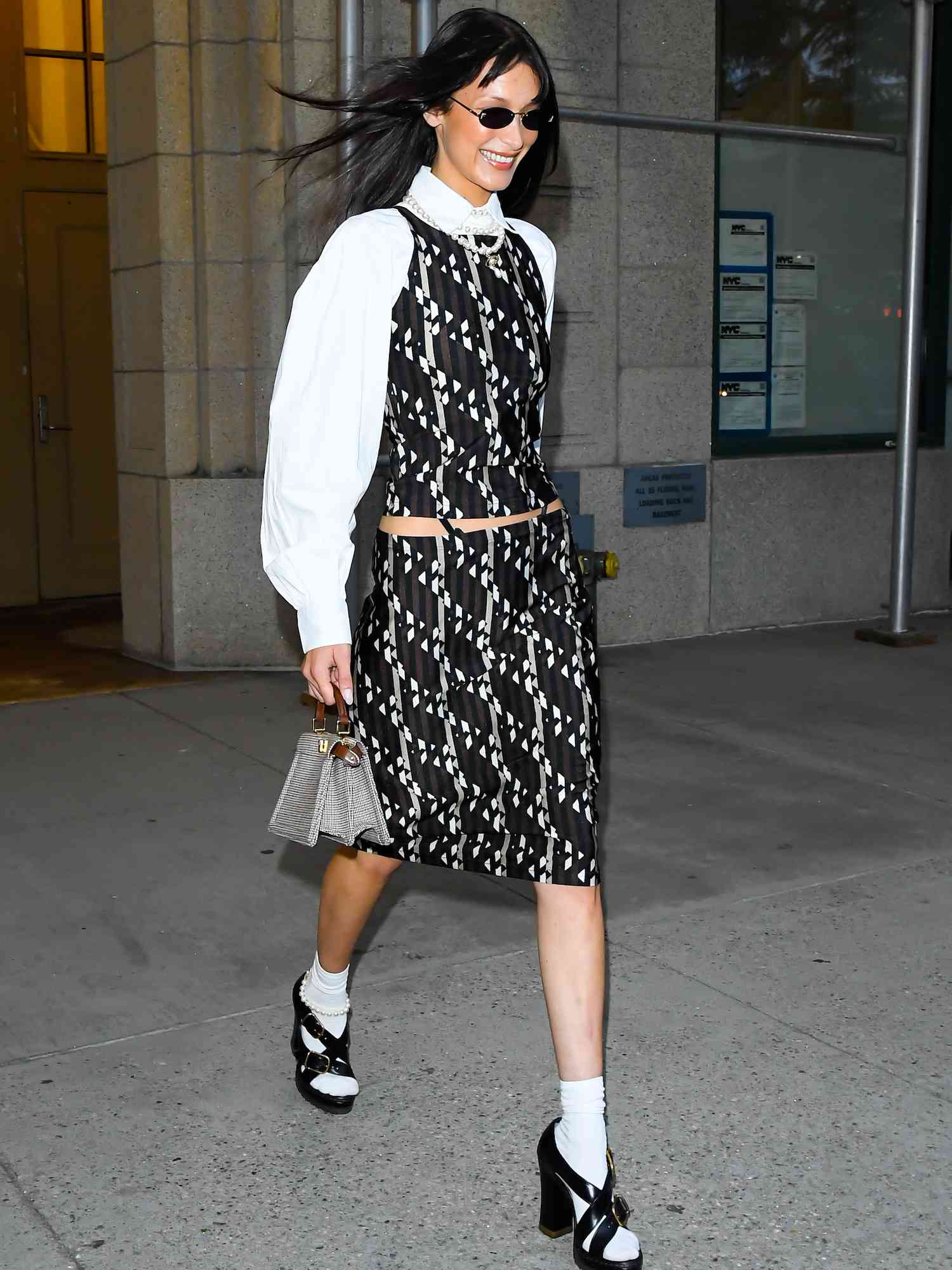 Bella Hadid em conjunto de top e saia preto e branco, blusa branca, óculos escuros minúsculos e sapatos plataforma