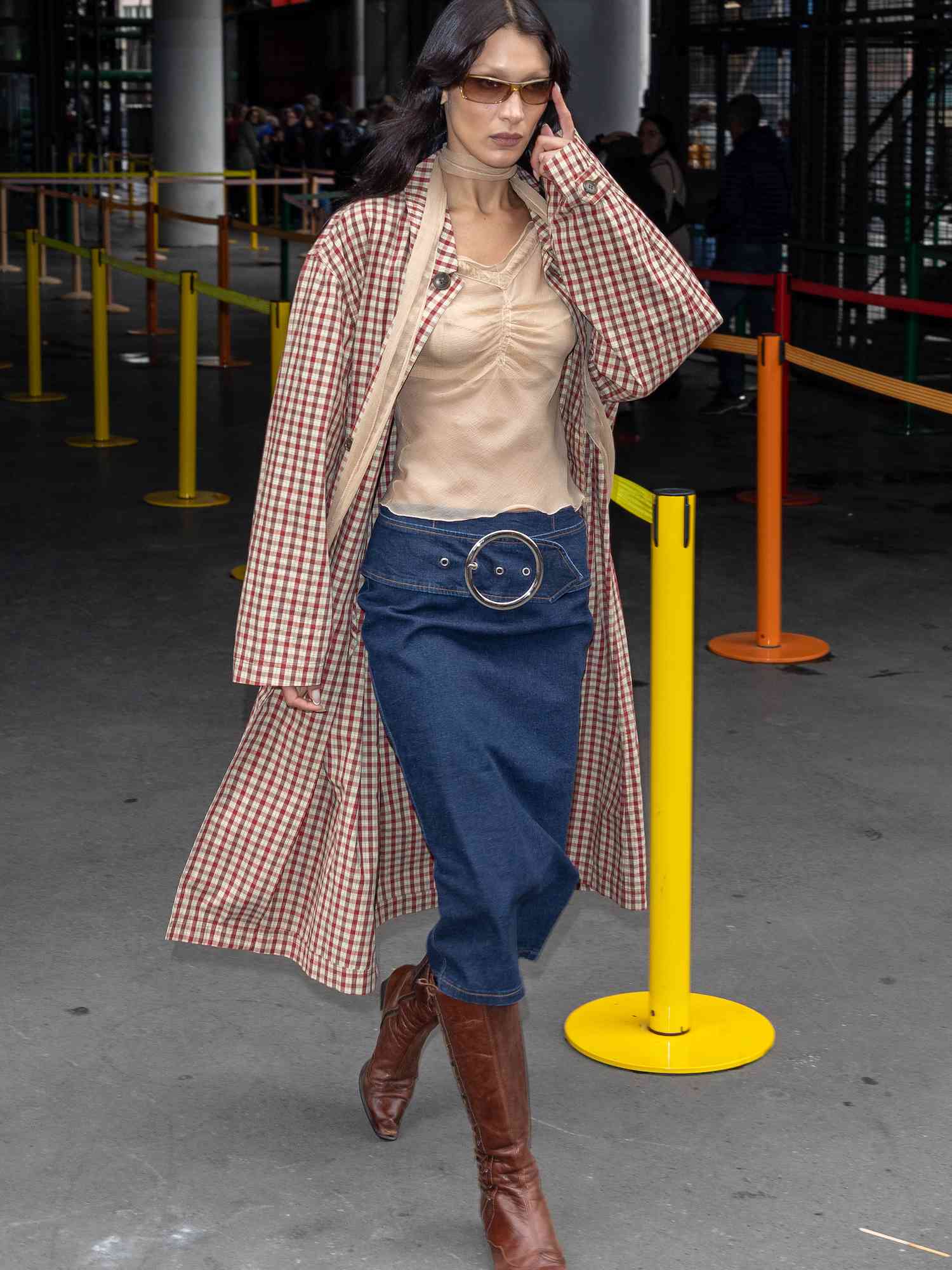 Bella Hadid com blusa nude, saia midi jeans, botas de couro marrom, casaco de algodão e lenço justo