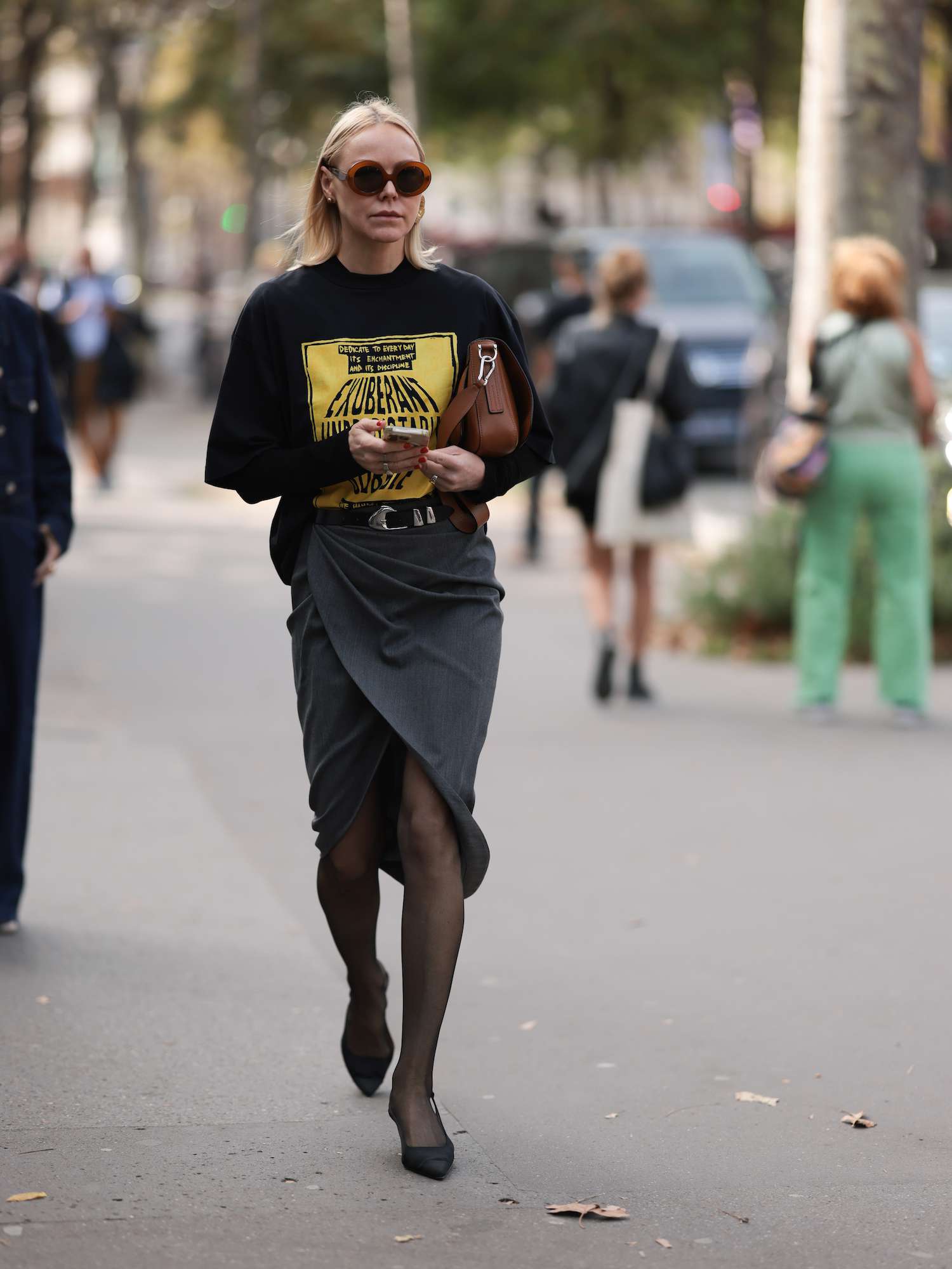 Mulher em uma camiseta gráfica preta, meia-calça cinzenta e transparente, sapatos pretos, sacolas marrons e óculos de sol marrons