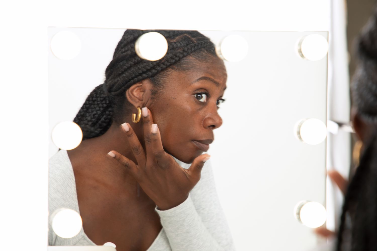 Uma mulher aplica um creme tonal Huda Beauty Glelowish Multidew Vegan Skin Tint Foundation em seu rosto, olhando no espelho